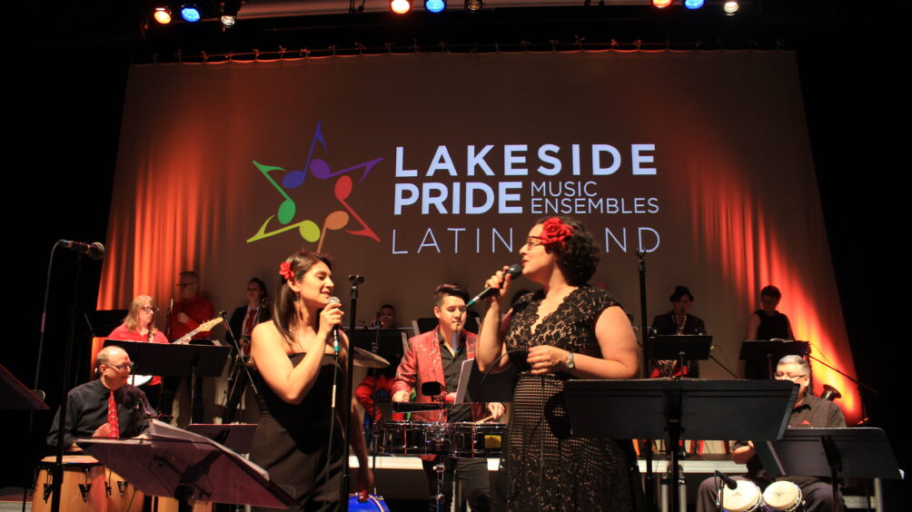 Latin Band performance with two singers front and the band in the back. Behind the band is a red curtain with the Lakeside Pride Latin Band logo projected.